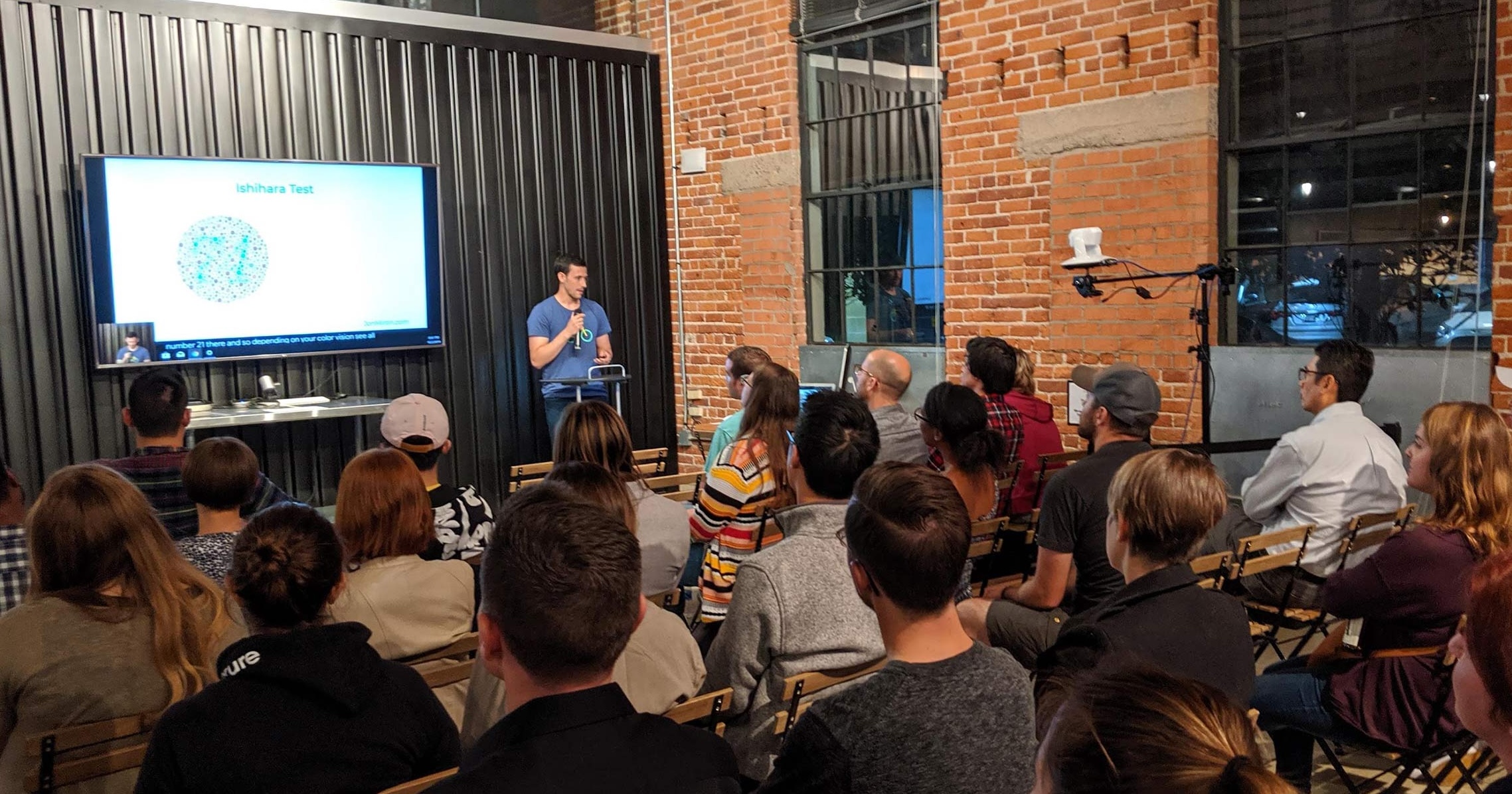 Jon Miron speaking to a seated audience, with an Ishihara colour-vision test slide on the screen behind him.